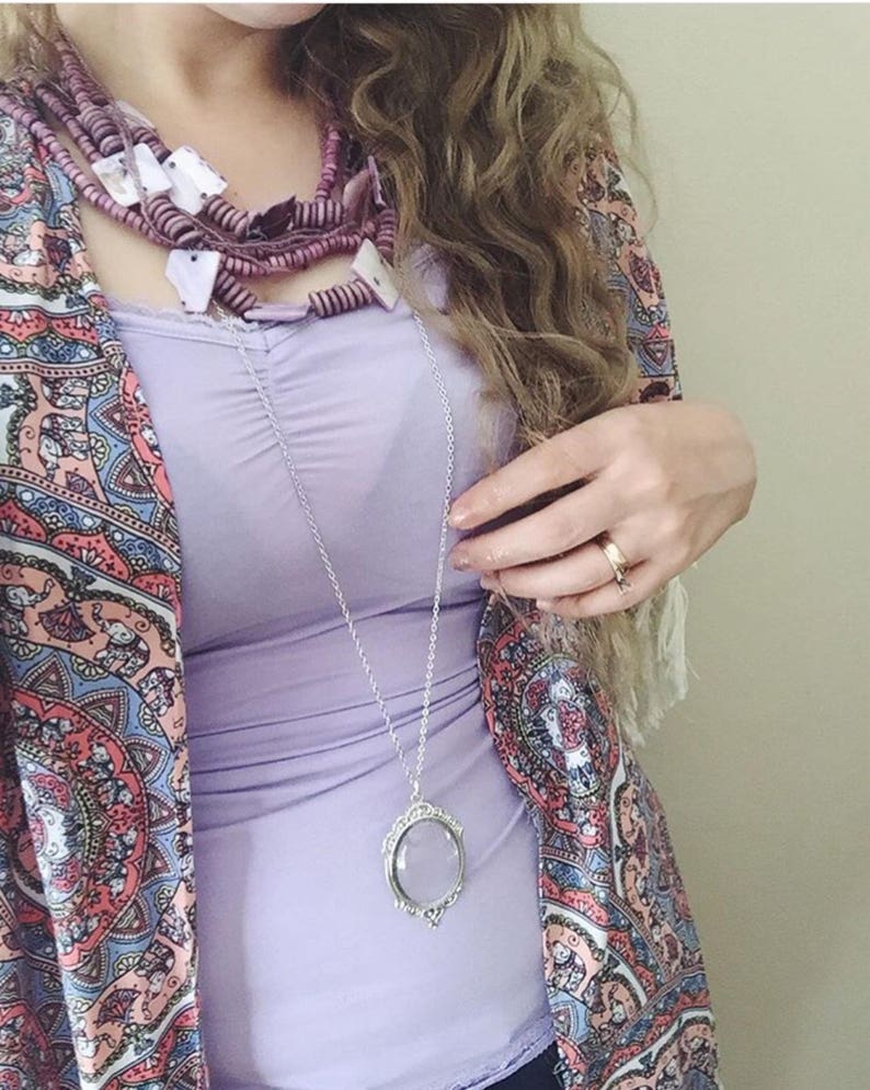 May include: A woman wearing a purple tank top with a layered necklace featuring a magnifying glass pendant. She is also wearing a colorful paisley patterned kimono and a ring on her finger.