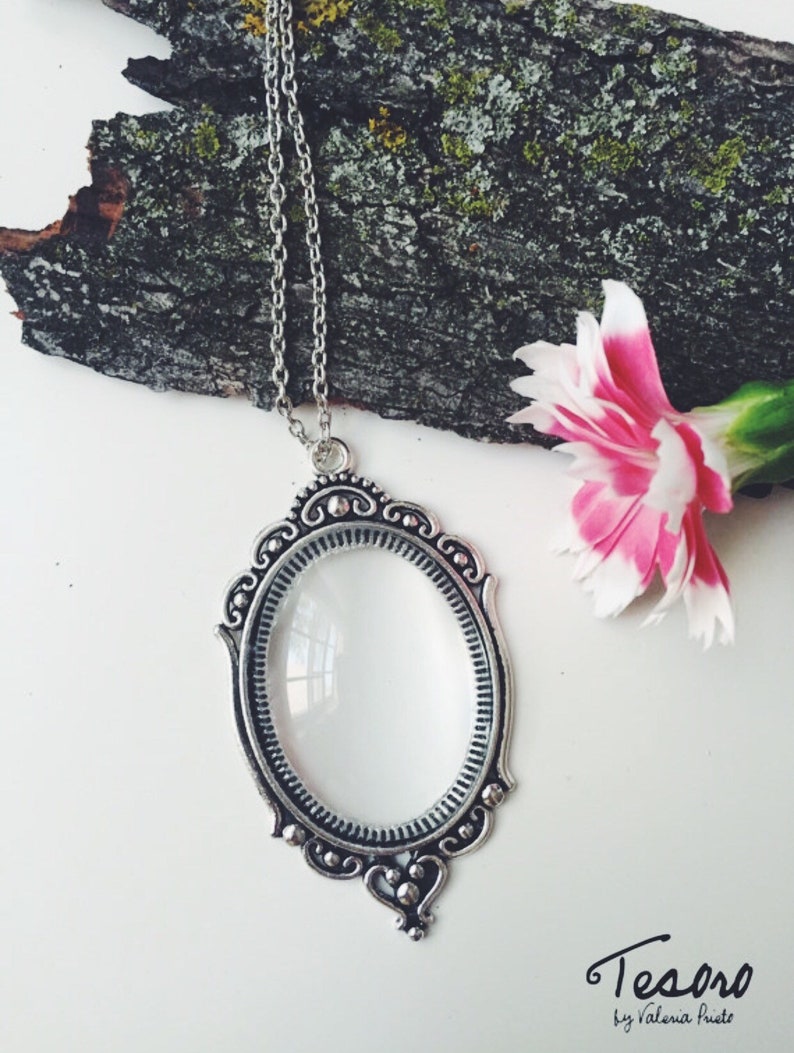 May include: A silver necklace with a magnifying glass pendant. The pendant is oval shaped and has a decorative frame. The necklace is on a white background with a pink flower.