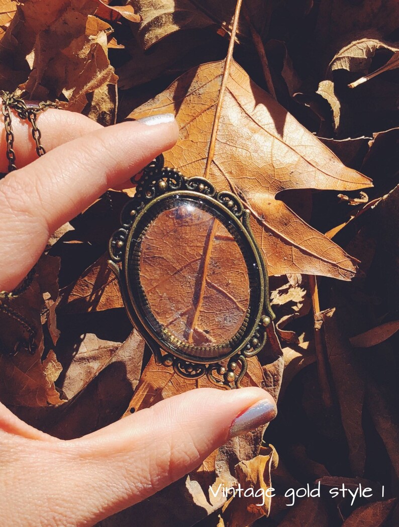 May include: A vintage gold-toned pendant necklace with a clear glass oval charm. The charm is filled with a dried leaf, and the necklace is lying on a bed of brown leaves.