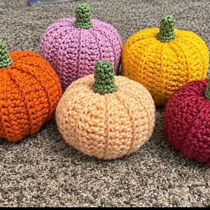 May include: A collection of five crocheted pumpkins in various colors including orange, light purple, yellow, peach, and dark red. Each pumpkin features a textured, ribbed surface and a small, green crocheted stem attached to the top center.