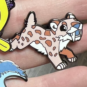 Puede incluir: Cuatro pines de esmalte con criaturas prehistóricas de dibujos animados. Los pines incluyen un dinosaurio azul y morado, una criatura amarilla con forma de pez, un tigre dientes de sable y un dinosaurio azul con dientes naranjas. Cada pin tiene un contorno negro.