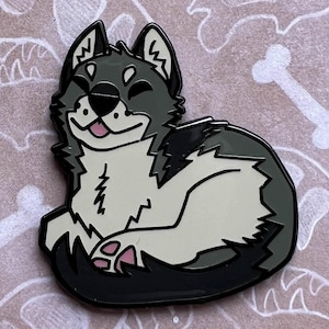May include: Paleopins enamel pin featuring a cartoon wolf with a black, white, and gray color scheme. The wolf is smiling and lying down. The pin is on a brown card with white dinosaur bones and the text "Paleopins" and "Andrea Radeck".