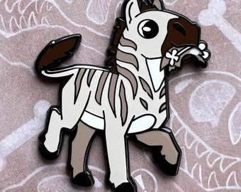 Könnte beinhalten: Paleopins-Emaille-Pin mit einem Cartoon-Zebra mit braunem und weißem Streifenmuster, das eine Blume im Maul hält. Der Pin befindet sich auf einer braunen Karte mit weißen Dinosaurierknochen und dem Text "Paleopins" und "Andrea Radeck".