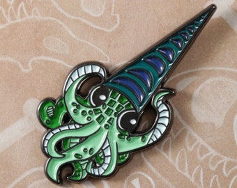 May include: Paleopins enamel pin featuring a green and blue cartoon squid with a pointed hat. The pin is on a brown background with white dinosaur bones.