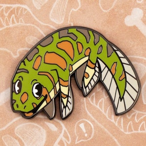 May include: Paleopins enamel pin featuring a green and orange prehistoric fish with a white fin. The pin is on a brown background with a white dinosaur bone design.