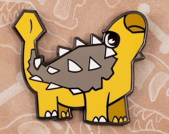 May include: A yellow and gray enamel pin featuring a cartoon Ankylosaurus dinosaur with the text "Paleopins" and "Andrea Radeck" on the background.