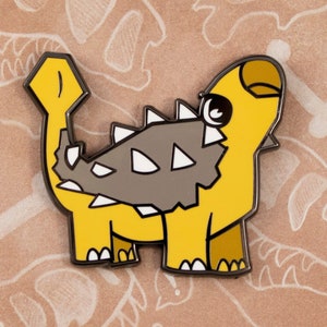 Może przedstawiać: Szpilka emaliowana w kolorze żółtym i szarym przedstawiająca kreskówkowego ankylozaura z tekstem "Paleopins" i "Andrea Radeck" na tle.