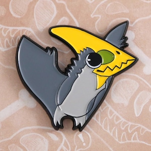 May include: A gray and yellow enamel pin of a cartoon pterosaur with big eyes. The pterosaur has a yellow crest and a gray body. The pin is on a brown background with a white dinosaur skeleton design.
