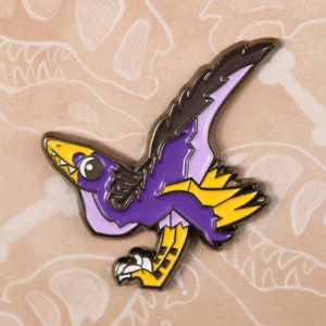 Może przedstawiać: Kolorowa emaliowana przypinka przedstawiająca stylizowanego dinozaura z fioletowymi, żółtymi i czarnymi akcentami. Przypinka umieszczona jest na beżowym tle z napisem "Paleopins" i ilustracjami szkieletów dinozaurów.