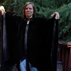 May include: A black velvet cape featuring a wide, flowing silhouette with a structured collar and a decorative black lace detail at the neckline. The garment is made of a soft, light-reflecting fabric that drapes over the shoulders and arms.