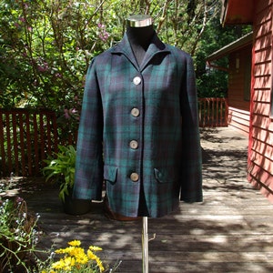 May include: A green and navy tartan jacket with four large, round buttons. The jacket features a classic collar and two front pockets. It is displayed on a mannequin against a backdrop of greenery and a wooden deck.