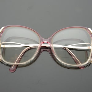 May include: Vintage oversized eyeglasses with a pink and gold frame. The glasses have a rounded shape and clear lenses. The temples and bridge are a light gold color. The frames are a translucent pink.