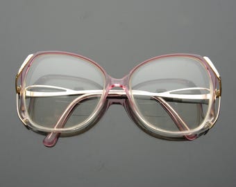 May include: Vintage oversized eyeglasses with a pink and gold frame. The glasses have a rounded shape and clear lenses. The temples and bridge are a light gold color. The frames are a translucent pink.