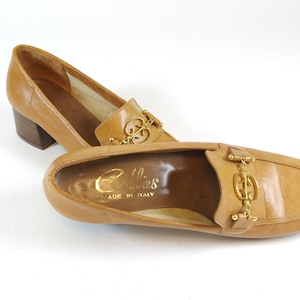 May include: A pair of brown leather heeled loafers with a gold chain and buckle detail. The shoes are made in Italy by the brand "Collies".