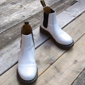Könnte beinhalten: Zwei weiße Chelsea-Stiefel aus Leder mit schwarzen elastischen Seiteneinsätzen und gelben Nähten um die Sohlen. Die Stiefel stehen auf einer verwitterten Holzoberfläche. Der Markenname ist auf der Zuglasche sichtbar.