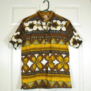 May include: Short-sleeved button-down shirt with a vibrant floral and geometric pattern in shades of brown, white, yellow, and green. The shirt is hanging on a green hanger against a white door. The shirt has a classic collar and button closure.