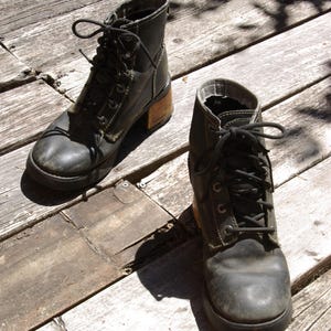 May include: A pair of black leather ankle boots with laces, featuring a chunky sole and a wooden heel. The boots are worn and have visible stitching details. The boots are placed on a weathered wooden surface.