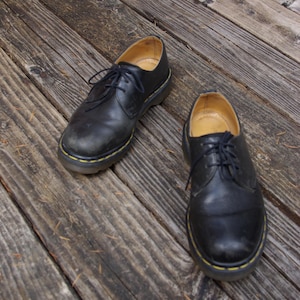 May include: A pair of black leather Oxford shoes with yellow stitching around the soles. The shoes have laces and are placed on a weathered wooden surface. The shoes appear worn.