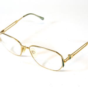 May include: Gold-tone eyeglasses with a rectangular frame and clear lenses. The temples and bridge are thin and delicate. The earpieces are green. The glasses are set against a white background.
