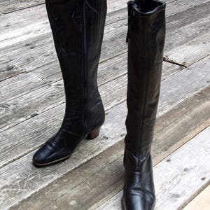 May include: A pair of black knee-high boots with side zippers. The boots have a slight heel and a rounded toe. The leather appears to be slightly worn, giving them a vintage look. The boots are displayed on a wooden deck.