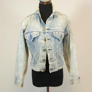 May include: Light blue denim jacket with a faded, acid wash effect. The jacket features a classic design with button closures, chest pockets, and a red logo tag. The jacket is displayed on a mannequin.
