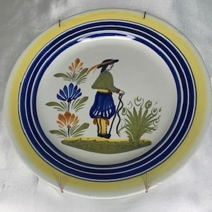 May include: Decorative ceramic plate with a hand-painted design. The plate features a central image of a figure in traditional attire, surrounded by floral motifs. The rim has concentric bands of yellow and blue. The plate is held by a gold-colored metal stand.
