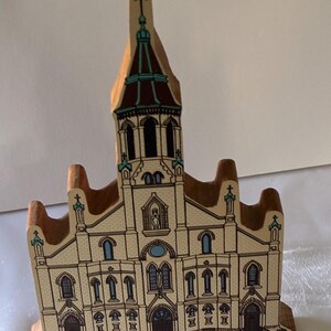 May include: A wooden cutout of a church building featuring a tall central steeple, multiple arched windows, and a detailed facade. The structure is painted in cream, brown, and teal tones, showcasing architectural elements like crosses and doorways.