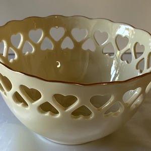 May include: A cream-colored ceramic bowl featuring a scalloped rim and a decorative openwork lattice pattern composed of repeating heart-shaped cutouts around the upper portion of the vessel, finished with a thin brown painted edge.