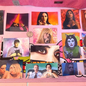 May include: A collection of colorful art prints featuring portraits of celebrities and pop culture figures. The prints vary in size and style, with some depicting close-ups and others full figures. The images are arranged on a pink surface.
