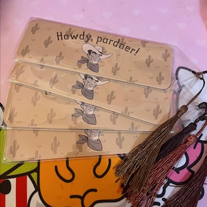 May include: Four beige bookmarks with a cowboy illustration and the text "Howdy, pardner!". Each bookmark features a cowboy with a hat and a cigarette. The background has cactus illustrations. Tassels in brown, black, and red are attached.