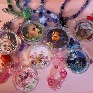 Puede incluir: Una colección de dijes circulares de acrílico transparente tipo shaker con varias ilustraciones de personajes y rellenos de confeti de colores. Cada dije está unido a un cordón de cuentas con cuentas de plástico variadas en formas de estrellas y corazones.
