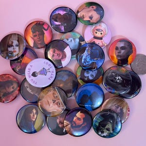 May include: A collection of circular pin-back buttons featuring various character illustrations and portraits, including one light purple button with the text YOU WEAR A TUTU, TOOT, arranged alongside a silver quarter for scale on a pink surface.