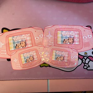 May include: Four pink Game Boy Advance game cartridges with a holographic finish. Each cartridge features a different Pokemon character on the front. The cartridges are arranged in a row, with the largest cartridge at the top and the smallest cartridge at the bottom.