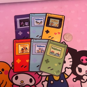 Op de afbeelding: Een verzameling kleurrijke, retro Pokémon Game Boy-stickers. Elke sticker heeft een ander kleurenschema, waaronder paars, blauw, geel, rood en groen, met een Pokémon-gamescherm en het woord "Pokémon".