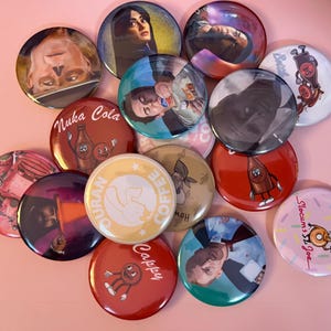 Könnte beinhalten: Eine Sammlung runder Ansteckbuttons mit verschiedenen grafischen Motiven, darunter Charakterporträts, Nuka-Cola-Branding, Slocum's-Joe-Donut-Grafiken, Cappy-Maskottchen-Illustrationen und Duran-Coffee-Logos in einer Vielzahl von Farben und Stilen.