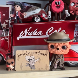 May include: A display of Fallout-themed collectables, featuring a red Nuka Cola truck and a figure wearing a cowboy hat. A sign with the text "Howdy, pardner!" is in front of the figure.
