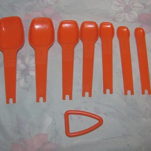 May include: A collection of orange measuring spoons and a small orange triangular tool. The spoons are of varying sizes, with long handles and a small notch at the base. The items are arranged on a floral patterned surface.