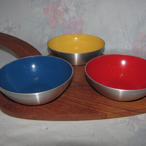 May include: A wooden serving tray with a handle and three metal bowls. The bowls are yellow, blue, and red.