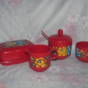 May include: A red vintage kitchen set featuring a butter dish, creamer, sugar bowl, and small bowl. Each piece is adorned with a floral design in yellow, blue, and green. The set is made of plastic.