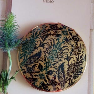 May include: A round compact mirror featuring a metallic gold finish with a detailed black and dark green floral paisley pattern, displayed alongside a small bundle of green spiky thistle flowers tied with a piece of brown twine.
