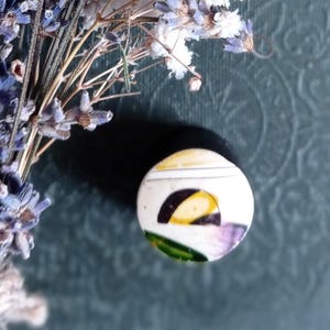 Può includere: Un piccolo bottone circolare in ceramica con base bianca e un design grafico composto da un semicerchio giallo, un arco nero e una striscia verde, posizionato accanto a un mazzetto di lavanda viola essiccata e fiori bianchi di gipsofila.