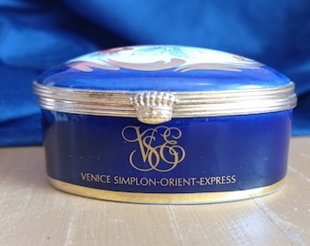 May include: A blue and gold porcelain trinket box with a lid. The box has a gold inscription that reads "Venice Simplon-Orient-Express".