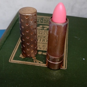 Peut inclure: Un rouge à lèvres vintage avec une mine rose, dans un étui de couleur bronze. L'étui présente un motif d'étoiles. Le rouge à lèvres est ouvert, révélant la couleur rose. Le capuchon est à côté du rouge à lèvres. Le rouge à lèvres est sur un livre vert.