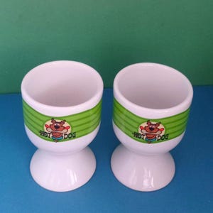 May include: Two white ceramic egg cups with a green band featuring a cartoon hot dog character. The egg cups have a flared base and a cylindrical top, ideal for serving soft-boiled eggs.