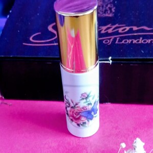 Puede incluir: Un frasco de perfume blanco con tapa dorada y diseño floral con rosas rosas y una mariposa azul. El frasco está sobre una superficie rosa con lavanda seca y flores blancas.