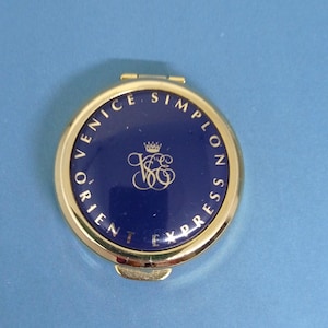 May include: A gold-toned, round compact mirror with a navy blue center. The text "VENICE SIMPLON ORIENT EXPRESS" is printed around the edge in gold. A gold emblem is in the center. The compact is on a light blue surface.