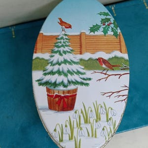 May include: An oval tin featuring a winter scene with a snow-covered Christmas tree in a wooden pot tied with a red ribbon, a fence, and two red robins. The tin has a silver trim and is set on a blue surface.