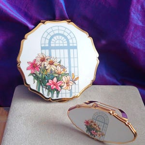 Peut inclure: Deux poudriers décoratifs. Les miroirs blancs à bord festonné présentent une bordure dorée et un motif floral avec une fenêtre. Un miroir est ouvert et l'autre est fermé avec une poignée violette.
