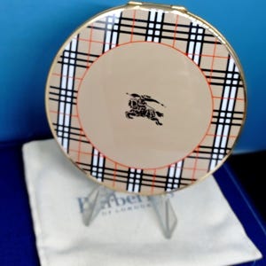 May include: A round compact with a beige center featuring a black equestrian logo. The outer rim has a classic plaid pattern in black, red, and white. It rests on a clear stand, above a white cloth and a blue box.