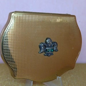 May include: A gold-toned compact with a textured surface and a decorative emblem featuring green and clear accents. The compact is displayed on a clear stand against a gold glitter background.